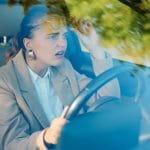 A young woman in a khaki suit pushes up her glasses and looks surprised as she drives a leather-upholstered car.