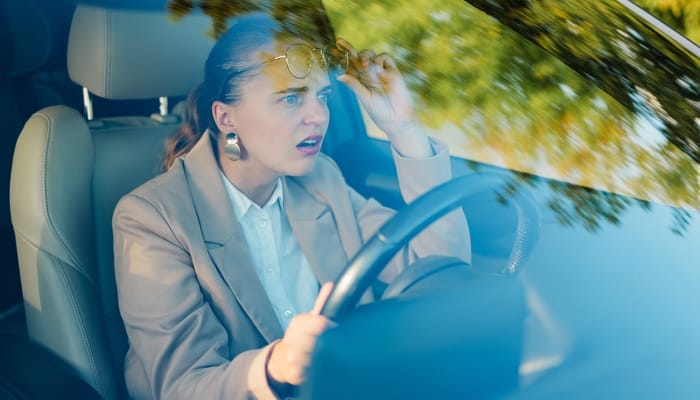 A young woman in a khaki suit pushes up her glasses and looks surprised as she drives a leather-upholstered car.