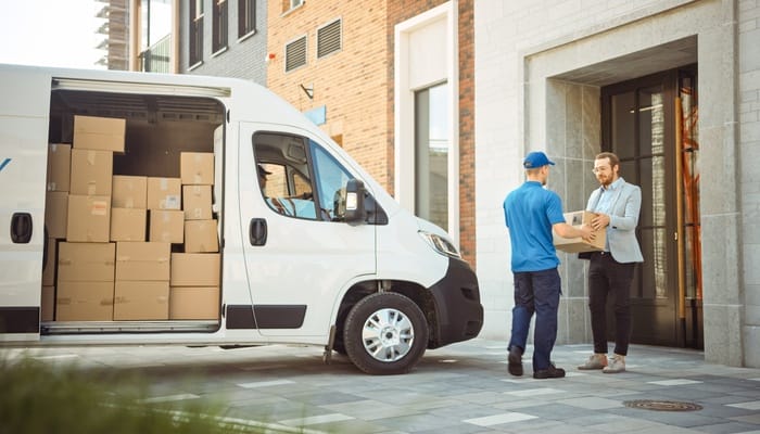 A white cargo van parked next to two people talking to each other. The van door is open with packages stacked up.