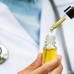 A doctor with a stethoscope holding an eye dropper full of CBD oil. They are holding it above a small bottle full of it as well.