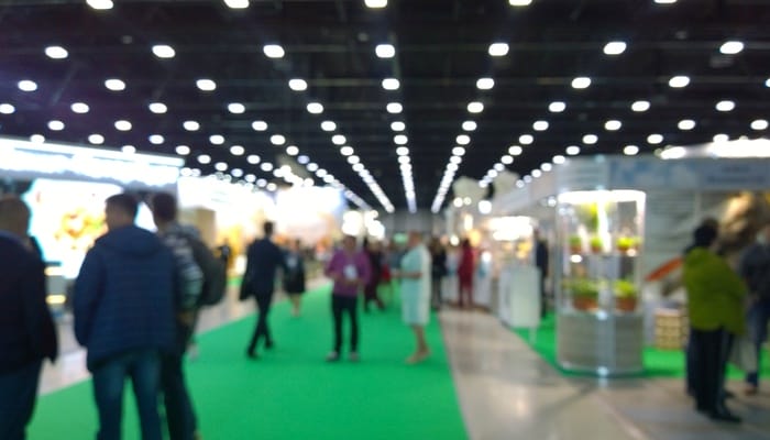 A blurry view of a convention center event with green carpeting in the aisles and booths on set up around the space.