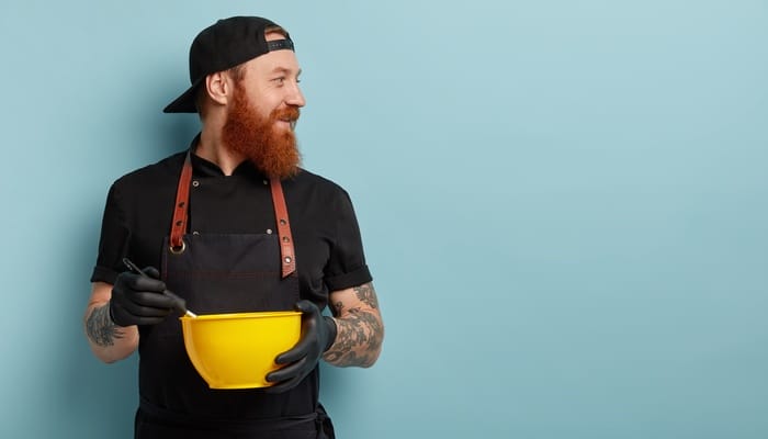 A man with a ginger beard and sleeve tattoos on both arms wears a baking apron and holds a mixing bowl.